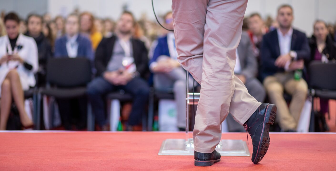 Relatore su palco rivestito con pavimento tessile rosso durante un congresso, con pubblico seduto sullo sfondo.