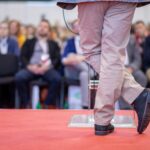 Relatore su palco rivestito con pavimento tessile rosso durante un congresso, con pubblico seduto sullo sfondo.