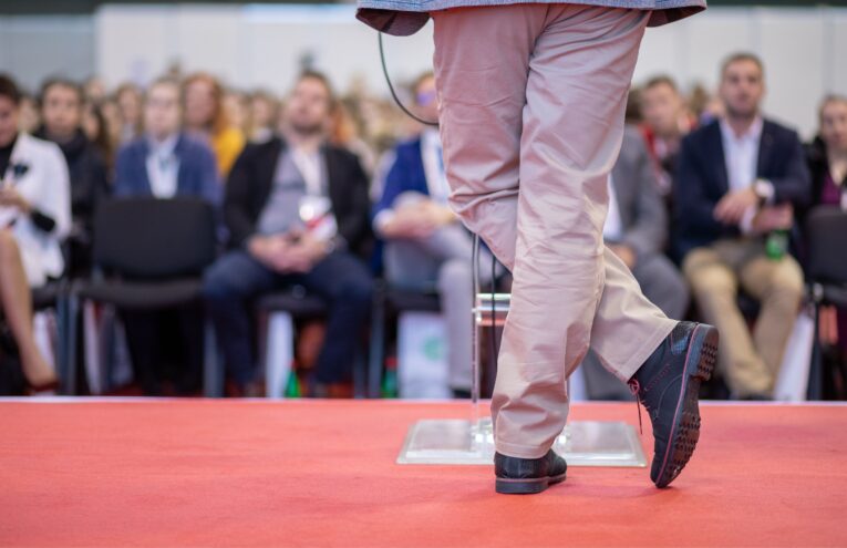 Relatore su palco rivestito con pavimento tessile rosso durante un congresso, con pubblico seduto sullo sfondo.